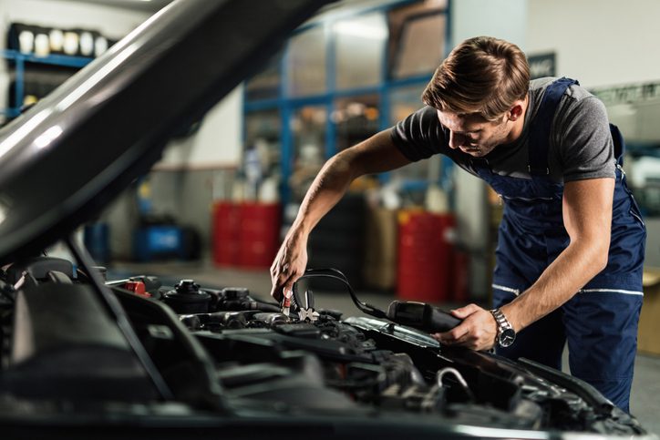 young mechanic attaching jumper cables on car engine while working in auto repair shop.